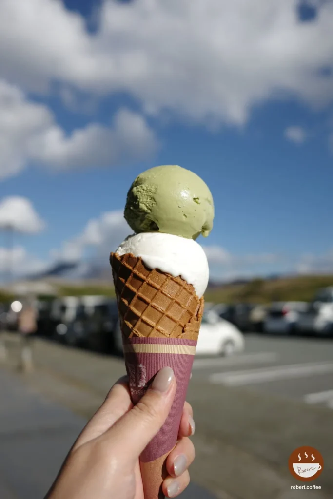 日本 熊本縣阿蘇市 ニュー草千里 ASO MILK ice cream shop：抹茶加牛奶的 ASO MILK 雙球甜筒，背景是藍天和停車場
